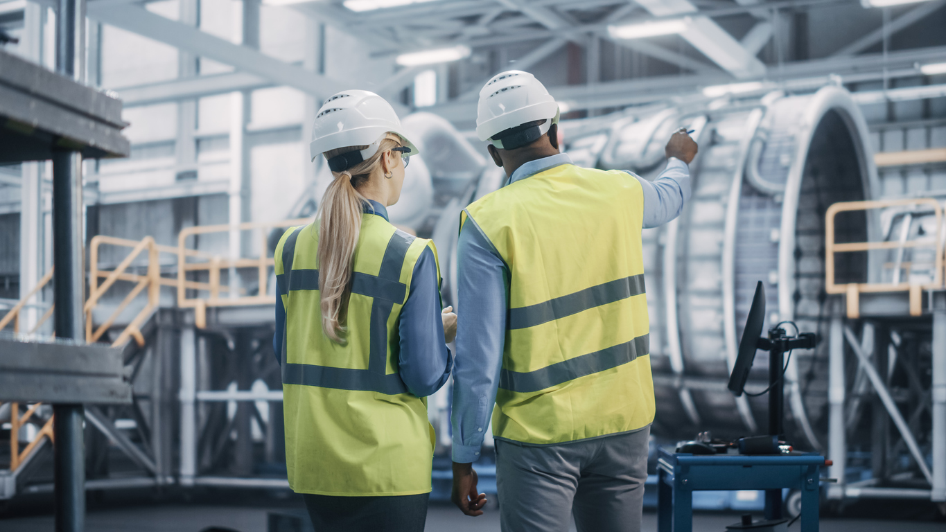 Two Diverse Professional Heavy Industry Engineers Wearing Safety Uniform and Hard Hats Working on Laptop Computer. African American Technician and Female Worker Talking on a Meeting in a Factory. IT specialisms
